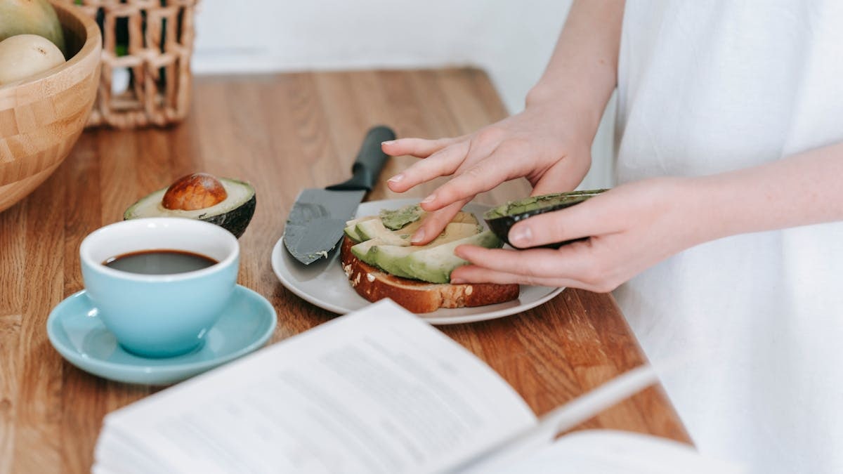 Crop anonymous female in white t shirt preparing toast with sliced avocado and cup of coffee for breakfast at wooden table